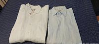Two men's medium size shirts: white guayabera on left and light blue dress shirt on right, folded on dark blue fabric.