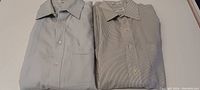 Two folded men's long sleeve dress shirts, one light blue and one beige/tan with stripes, on a flat surface.