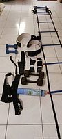 Photo of sport equipment including two pairs of dumbbells, adjustable weights, a black harness, agility ladder, and a yoga mat arranged on a tiled floor.