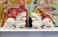 Photo of two white stone foo lion statues side by side in front of colorful abstract paintings.
