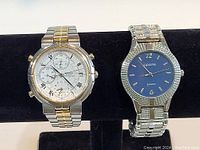 Two men's wristwatches displayed on stand, one Seiko chronograph with white dial and two-tone bracelet, other Geneva quartz with blue dial and textured silver-tone band.