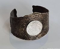 Front view of antique-looking bangle bracelet with textured dark metal surface and a silver-colored coin featuring Arabic writing and star design.