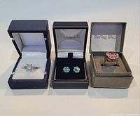 Three jewelry items displayed in individual boxes: two rings and one pair of earrings. The ring on the left has a clear square stone, the pair of earrings in the center are turquoise with silver inlay, the ring on the right has a large rectangular pink stone.