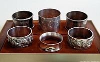 Set of six silver napkin ring holders placed on a brown felt-like tray, showing various embossed decorative designs and polished finish.
