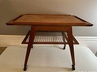 Full front view of the teak TV/bar trolley showing tray top, tapered legs, mesh lower shelf, and casters.