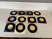 Top down view of 12 vintage 7-inch vinyl records with original sleeves, arranged on a white floor showing front labels and sleeves.