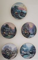 Five decorative plates with winter cabin scenes shown hanging on a wall, each 8.5 inches round with visible wire hangers.