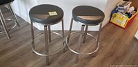 Two grey and chrome bar stools with round seats and chrome legs with footrests, placed on wood floor.