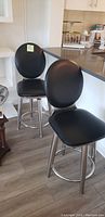 Photo showing two black bar stools with padded round backs and seats, stainless steel bases at counter height beside kitchen counter.
