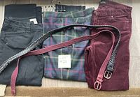 Photo showing three pairs of ladies pants: black slacks, burgundy corduroy pants, and plaid capris with two belts on top of the pants and a carpet background.