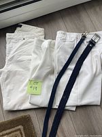 Three white women's pants stacked, two navy woven belts with silver buckles laid over.