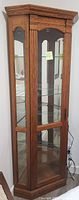 Full view of the oak corner display cabinet showing its glass sides, front door, wooden frame, and four internal glass shelves.