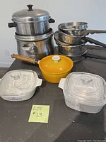 Full lot overview showing two Corningware casseroles, Descoware yellow pot, and stainless steel pots and pans.
