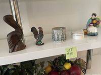 Wide view showing all items on white shelf: wooden pelican, rooster figurine, candle holder, white round Pandora jewelry box, and ceramic balloon lady.