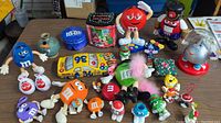 Overview of assorted M&M character toys and memorabilia including figurines in various colors, a holiday-themed M&M container, and a toy car.