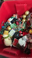 Red plastic tote filled with assorted Christmas ornaments, including baubles, a penguin plush, Santa Claus figure, and string lights.