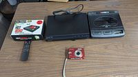 Top-down view showing all items together: the Roku Hot Rodge box, Sony DVD player, Amico game controller, red Polaroid camera, and a remote control on a wooden table.