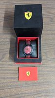 Ferrari watch resting in a black box with Ferrari logo on the lid, accompanied by a red informational card with Ferrari crest.
