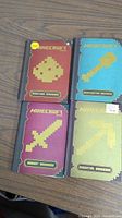 Four Minecraft books placed on a wooden table: Crafting Handbook, Combat Handbook, Building Handbook, Essential Handbook. Each has pixel art cover relating to Minecraft items.