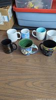 Seven assorted ceramic mugs on wooden surface with boxes in background.