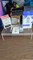 Lot items on white wire cabinet shelf on wooden table, includes boxed Honest shampoo & body wash, Honest body wash bottle box, Hunter boot socks in packaging, and small black cleaning brush.