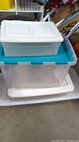 Three stacked plastic storage totes: clear with white lid, clear with teal lid, and large clear base with white lid underneath.