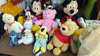 Front view of all plush toys showing Mickey Mouse, Minnie Mouse, Winnie the Pooh, Pluto, pink teddy bear, and other Disney and non-Disney stuffed animals.