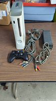 Full view of white Xbox 360 console with black controller, power supply and AV cables visible on wooden table.