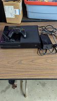 Xbox One console with controller on top and power supply cable to the side on a table