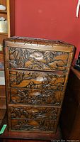 Front view of the carved wooden cabinet showing all five drawer fronts with bird and floral reliefs against a red wall.