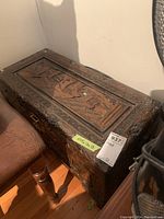 Angled side view showing the entire chest and its carved sides, positioned next to a chair and wall.