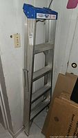 Full view of folded aluminum ladder with blue tool tray on top, leaning against wall near outlet and boxes.