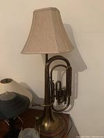 Brass wind instrument lamp mounted on wooden base with beige fabric lampshade, shown front angle.