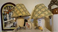 Two cream-colored lamps with leaves and orange floral patterned shades beside a round floral decanter and two metallic night lights on a white appliance.
