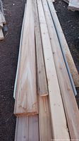 Pile of long pine and cedar lumber including shiplap and tongue & groove boards, outdoors on dirt ground.