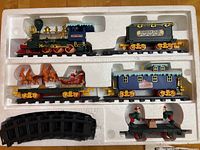 Full Christmas train set in original styrofoam packaging showing locomotive, coal car, sleigh car, passenger car, elf figure flatbed, and curved track pieces.