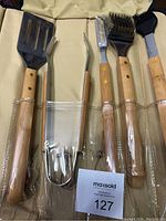 Photo of 5 BBQ utensils in beige carrying case, showing wooden handles and metal parts, sealed in plastic pockets.