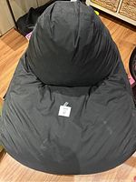 Black bean bag chair placed on hardwood floor with visible wrinkles and tag indicating lot number.