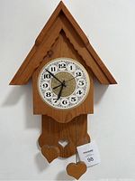 Front view showing the homemade pine birdhouse style clock with white decorative clock face, black hands and three heart-shaped wooden pendants hanging by chains from the bottom.