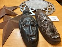 Two carved wooden tribal masks and two rust-colored 3D metal stars on a wooden surface with a silver round decorative wall piece in the background
