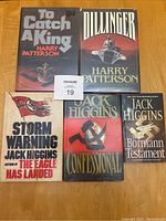 Lot of 5 Jack Higgins novels including To Catch a King, Dillinger, Storm Warning, Confessional, and The Bormann Testament shown on a wooden floor.