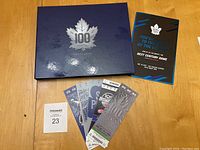 Closed booklet with silver embossed 100 Maple Leafs logo and several sample tickets and invitation card laid out on a wooden surface.