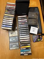 Photo showing a collection of cassette tapes stacked and spread on a wooden surface with a black TDK soft carrying case and black zippered case.