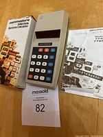 Commodore 776M calculator with original box and instruction manual laid out on table. Calculator body is beige with multicolored buttons and a dark red LED display. Box shows minor edge and corner wear.