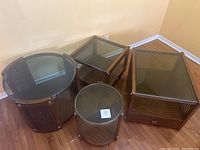 Overview of four wood and wicker coffee tables with glass tops in various shapes and sizes, placed on wooden floor near yellow walls.
