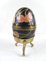 Front view of the cloisonné egg resting on its brass stand, showing pink floral decorations on dark blue background with brass base and trim.