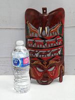 Full view of the African mask next to a water bottle to show scale. The mask is dark reddish-brown wood with carved animals and a human face.