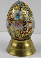 Front view of cloisonne egg with blue, white, lavender flowers and gold outlines on brass stand