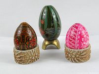 Three vintage decorative eggs displayed on stands: two ceramic textured bases, and one brass tripod base with floral and bird designs