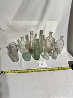 Full lot showing 11 varied antique glass bottles in clear, aqua and greenish tint, with measuring tape for scale
